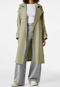 Olivově zelený trenčkot s klasickým límcem, dlouhými rukávy a bočními kapsami, doplněný šedými širokými dž�ínami a bílými teniskami.