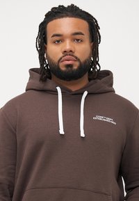 Hombre con barba y rastas, vistiendo una sudadera marrón con cordones blancos y pequeño texto blanco en el pecho, frente a un fondo liso.