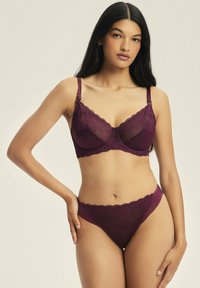 Koronkowy stanik i pasujące stringi w głębokim burgundzie. Posiada przezroczyste panele, falbaniaste krawędzie oraz regulowane ramiączka, co zapewnia pewne dopasowanie.
