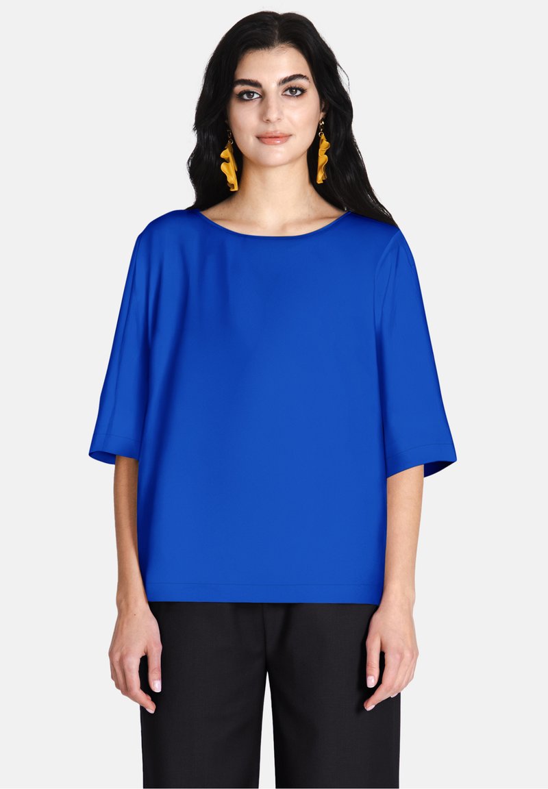 Top blu corto con maniche corte e scollatura ampia, dalla consistenza liscia. Indossato con pantaloni neri, caratterizzati da una vestibilità ampia e rilassata. Orecchini gialli visibili.