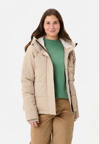 Giacca imbottita beige con cappuccio, dotata di zip frontale e tasche laterali. Indossata sopra una camicia verde e pantaloni marroni, con una trama trapuntata.