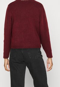 Maglione bordeaux in lana con scollatura a girocollo e orlo a coste, abbinato a jeans neri. La parte posteriore presenta sottili texture e dettagli di cucitura.