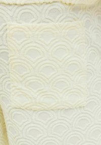 Tejido de color crema con un patrón de olas texturizado. Un pequeño bolsillo de parche con el mismo diseño es visible en la prenda.