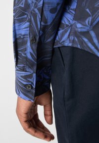 Gros plan d'une main près d'une poche portant une chemise à manches longues bleue et noire à motifs avec des poignets boutonnés et un pantalon sombre.