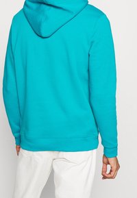 Sudadera con capucha turquesa con una capucha amplia, mangas largas y puños acanalados. Tela suave y texturizada con un ajuste relajado. Se ven pantalones blancos.