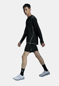 Sort langærmet skjorte med hvid syning, parret med sorte shorts. Model iført hvide sko og sorte sokker med logo. Enkel design.
