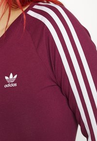 Burgundy sportstopp med vit tre-stripesdesign på ärmarna. Har en v-ringning och en liten vit Adidas-logotyp. Tyg i bomullsblandning.
