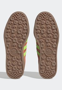 Ruskea kumipohja, jossa on teksturoitu pyöreä kuvio, ja jokaisella puolella kolme vihreää raitaa sekä adidas-logo kantapäässä.