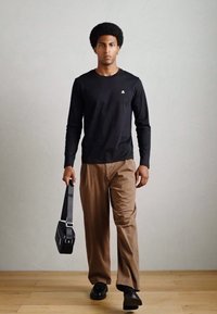 Cămașă neagră cu mâneci lungi, pantaloni maro lejeri, pantofi negri și o geantă de umăr neagră. Modelul stă pe un podea din lemn cu un fundal simplu.
