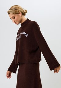 Ruskea fleece-neule, jossa on korkea kaulus, leveät hihat ja lyhyt leikkaus; edessä valkoinen teksti "OLD KARMA CLUB".