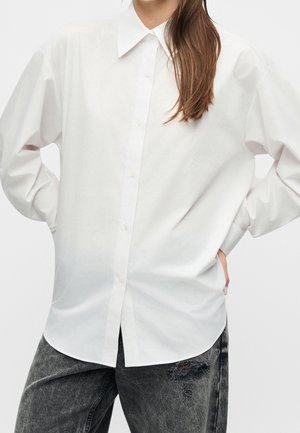 Kvinde iført en løs hvid knap-op-skjorte med spids krave og sorte acid-wash jeans med en lille rift på låret.