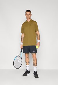 Nike polo skjorte i olivengrønn, kombinert med svarte shorts. Hvit Nike-logo, tennisracket, gul tennisball og svarte joggesko.