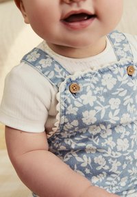 Lyseblå denimoveraller med hvidt blomstermønster, med træknapper og en blonderet halsudskæring. Bæres over en hvid ribbet top.