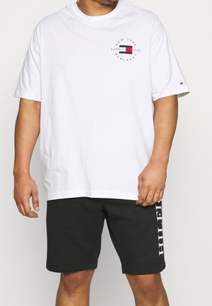 Homme portant un T-shirt blanc Tommy Hilfiger avec logo sur la poitrine et la manche, associé à un short noir avec texte blanc sur la jambe gauche.