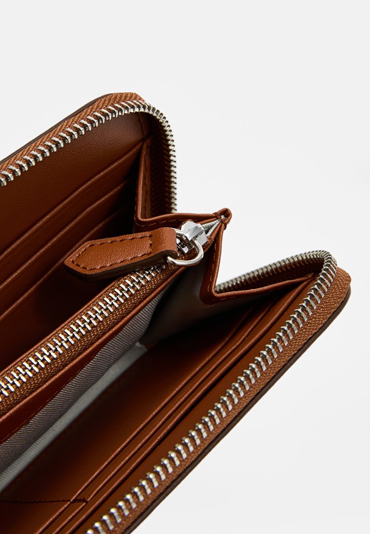 Portefeuille en cuir marron avec fermeture éclair, plusieurs emplacements pour cartes et un intérieur lisse. Doté de détails de fermeture éclair et de coutures en ton argent.