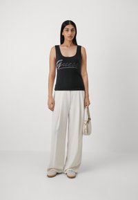 Canotta nera con scollatura a giro e logo "Guess" in argento scritto in corsivo. Abbinata a pantaloni larghi color crema e scarpe chiare.