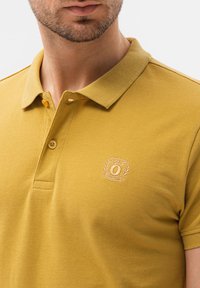 Polo shirt giallo senape realizzato in cotone texture, con una chiusura a due bottoni e logo ricamato sul lato sinistro del petto.