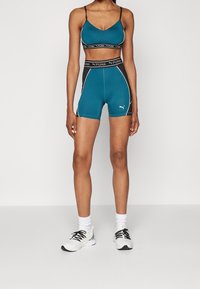 Kobieta nosząca turkusowy i czarny stanik sportowy Puma oraz dopasowane szorty, w białych butach sportowych i skarpetkach, stojąca na jednolitym tle.