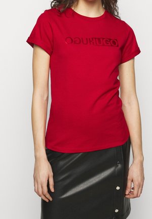 Kobieta ubrana w czerwoną koszulkę marki Hugo oraz czarną skórzaną spódnicę z srebrnymi guzikami po bokach.