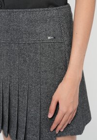 Grå plissert skjørt laget av teksturert stoff, med fiskebensmønster og en liten merkevarelogo på midjebåndet.