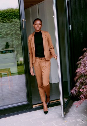 Costume marron avec une coupe ajustée, col roulé noir en dessous, et talons noirs. Se tenant devant une porte en verre avec un sol en pierre texturée.