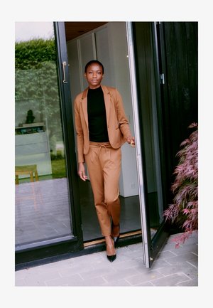 Costume marron avec une coupe ajustée, col roulé noir en dessous, et talons noirs. Se tenant devant une porte en verre avec un sol en pierre texturée.