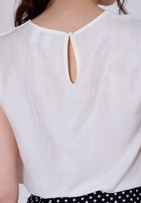 Blouse blanche avec une ouverture en goutte d'eau dans le dos, dotée d'une texture douce et d'un col rond, présentée avec une jupe à pois.