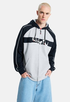 Junger Mann mit kurzen blonden Haaren, trägt einen grau-schwarzen Kapuzenpullover mit Reißverschluss und hellblaue Jeans, Hände in den Taschen, nach vorne blickend.