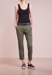 Top sans manches noir, pantalon cargo vert avec plusieurs poches et une ceinture noire, associé à des chaussures à enfiler à imprimé animal.