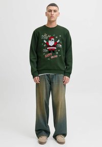 Sudadera verde con un gráfico de Santa con gafas de sol, rodeada de copos de nieve y texto. Combinada con jeans de denim sueltos y calzado beige.