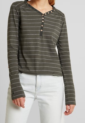 Femme portant une chemise à manches longues vert olive avec de fines rayures blanches et des boutons le long d'un large col, associée à un jean de couleur claire.