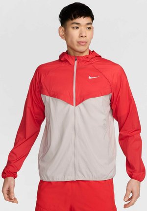 Veste légère rouge et grise avec fermeture éclair à l'avant, capuche et poignets élastiques. Présente le logo Nike sur la poitrine et l'épaule. Tissu lisse et texturé.