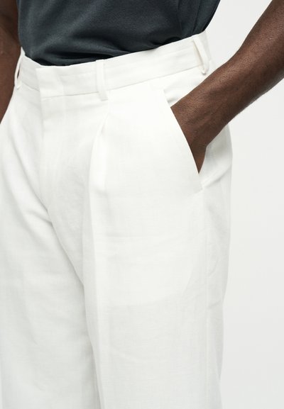 Personne à la peau foncée portant un pantalon blanc taille haute avec la main dans la poche latérale et une chemise gris foncé rentrée.