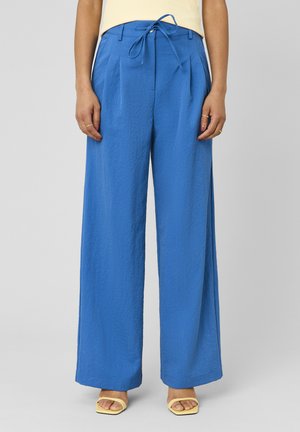 Femme portant un pantalon bleu taille haute, jambes larges avec plis sur le devant et ceinture à la taille, associé à des sandales jaunes à bout ouvert sur un fond uni.