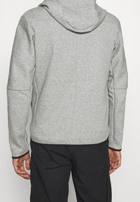 Šedá mikina s kapucí z texturované látky, vybavená kapucí a dlouhými rukávy. Na zádech jednoduchý design bez viditelných log nebo ozdob.
