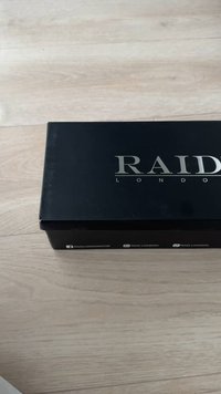 Caja negra con "RAID LONDON" en letras plateadas. Textura suave, forma rectangular y logotipos en los lados. Fondo de textura de madera.
