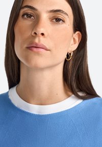 Pullover blu con un design a maglia testurizzata, caratterizzato da un colletto a coste bianco. Il modello ha capelli lunghi e scuri e indossa piccoli orecchini a cerchio.
