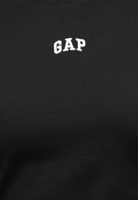 Μαύρο ύφασμα με λευκό κεντητό κείμενο "GAP" στο κέντρο κοντά στο πάνω μέρος.