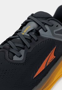 Zwarte hardloopschoen met een bovenwerk van mesh in grijs en oranje accenten, opvallende veters en een gestructureerde gele zool. "TORIN 8" branding zichtbaar.
