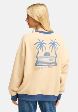 Blondynka w beżowej bluzie z niebieskimi wykończeniami oraz niebieską grafiką palmy i zachodu słońca na plecach, zestawiona z niebieskimi jeansami.