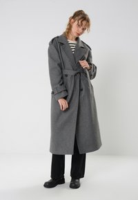 Cappotto trench grigio in lana con cintura in vita, bottoni doppi e ampi risvolti, abbinato a pantaloni neri e scarpe nere spesse.