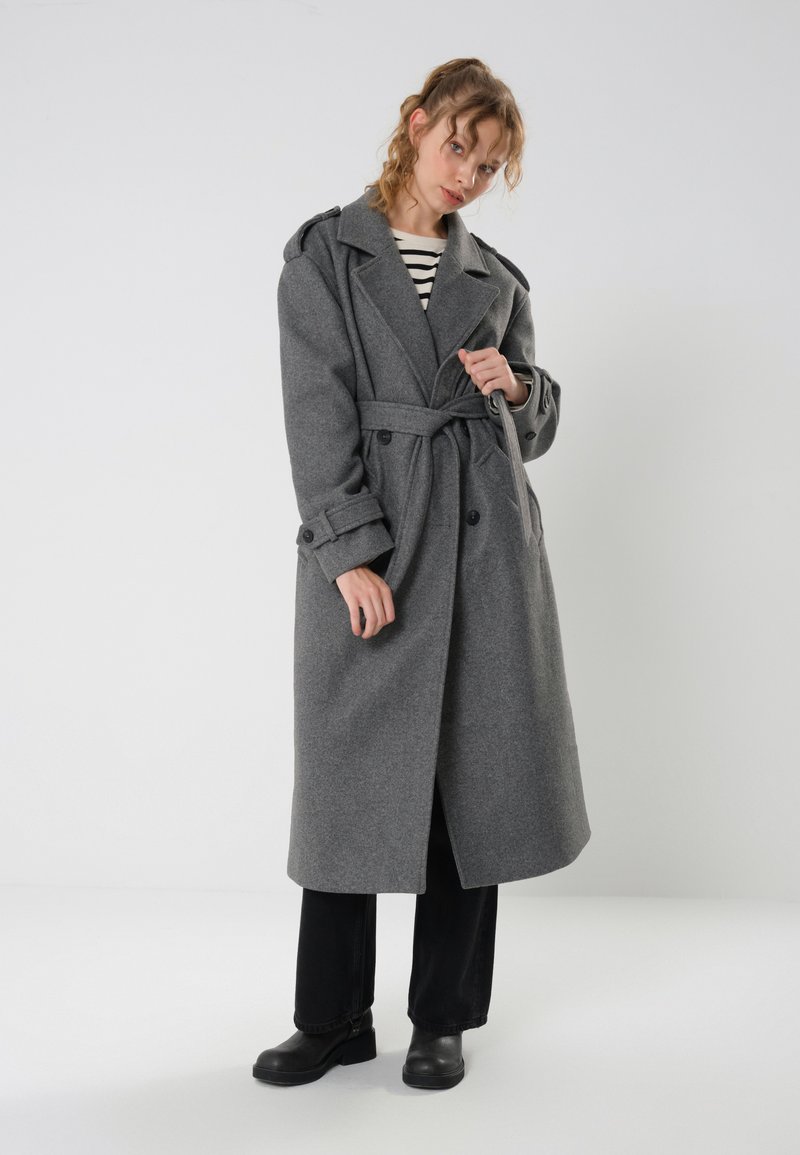 Cappotto trench grigio in lana con cintura in vita, bottoni doppi e ampi risvolti, abbinato a pantaloni neri e scarpe nere spesse.