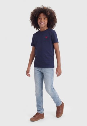 Dieťa s kučeravými vlasmi, oblečené v námorníckom modrom tričku, svetlom modrých džínsoch a hnedej obuvi, usmievajúce sa a stojace pred jednofarebným pozadím.