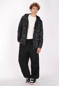 Giacca nera a camo con cappuccio, abbinata a pantaloni neri larghi e sneakers nere con dettagli bianchi e rossi. Tessuto dalla superficie liscia.