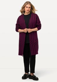 Cardigan viola con motivo a righe nere, design aperto sul davanti, maniche lunghe e vestibilità comoda, indossato sopra un top e pantaloni neri.