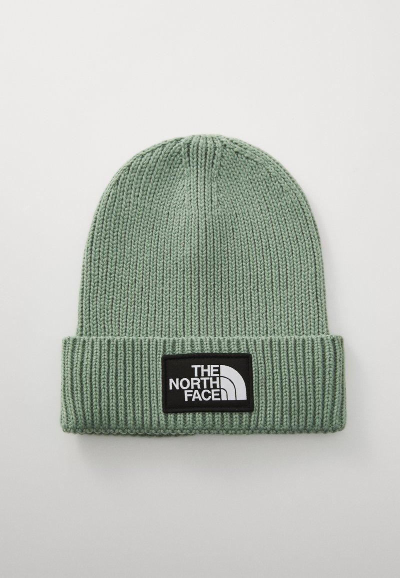 Gorro de malha em verde suave com uma aba dobrada, apresentando uma etiqueta preta com texto e logotipo em branco. Padrão canelado texturizado por toda a parte.