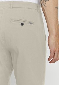 Beige Hose aus strukturiertem Stoff, mit einer Gesäßtasche mit einem einzelnen Knopf und einem dezenten gestreiften Akzent.