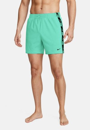 Uomo che indossa pantaloncini Nike turchesi con logo Nike nero e nastro con logo sul lato, in piedi a piedi nudi su sfondo bianco.