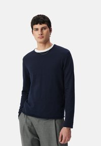 Granatowa koszula z długim rękawem, uszyta z miękkiego, strukturalnego materiału, z okrągłym dekoltem i białym wewnętrznym kołnierzykiem, zestawiona z szarymi, dopasowanymi spodniami.