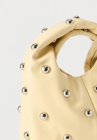 Κρεμ δερμάτινη τσάντα με κυκλικό χερούλι και ασημένιες μεταλλικές τρουκς ίσα κατανεμημένες στην επιφάνειά της, πάνω σε ανοιχτό φόντο.